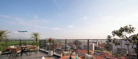 A terrace with outdoor seating, a view of the cityscape, and a plane in the distance.
