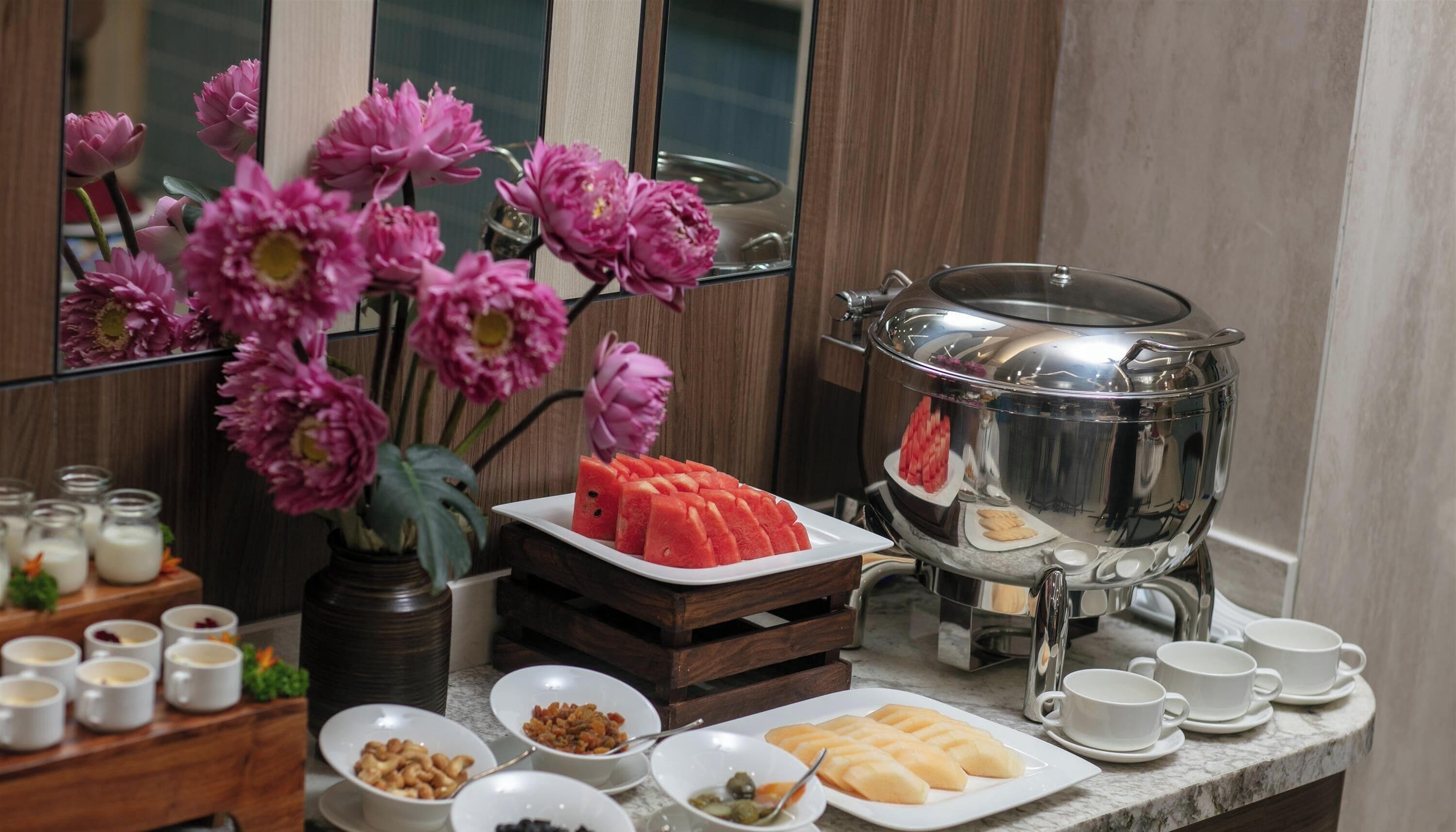 Montaje de desayuno buffet con diversos alimentos, una presentación de frutas y un jarrón decorativo con flores.