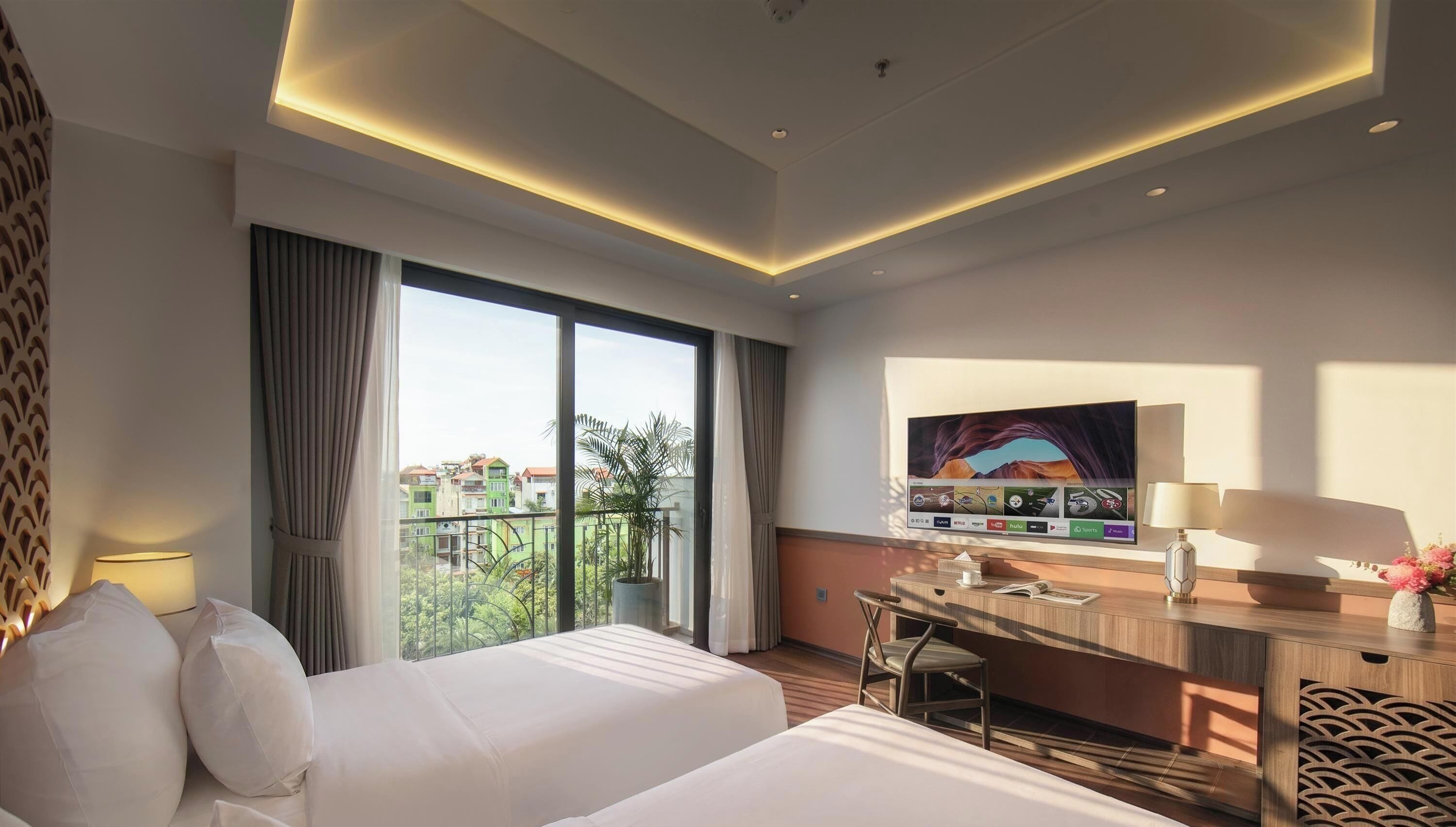 Une chambre d’hôtel équipée d’un lit, d’un bureau, d’une chaise et d’une grande fenêtre donnant sur des bâtiments et des espaces verts.