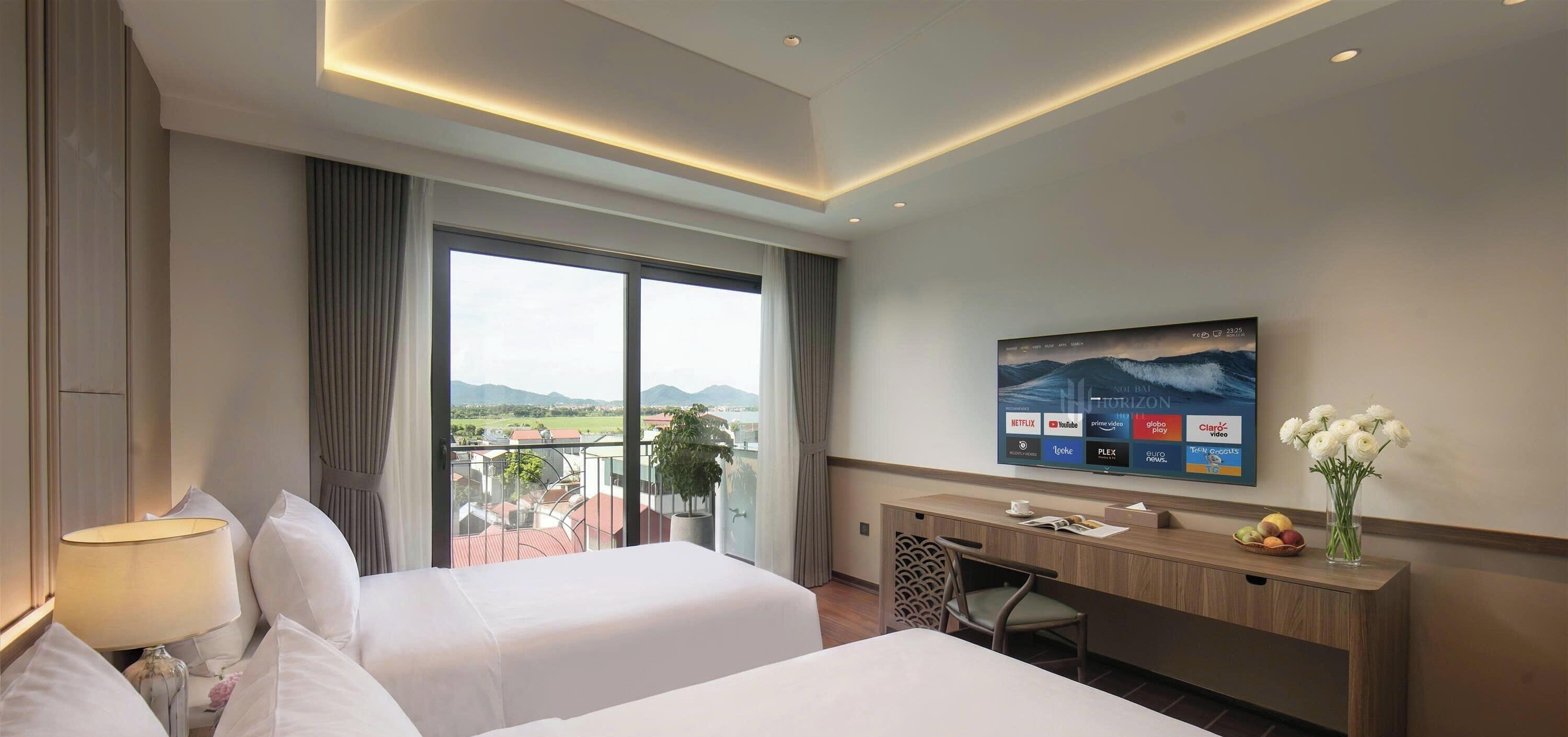 Habitación de hotel con dos camas, un escritorio, un televisor montado en la pared y un balcón con vistas al paisaje.
