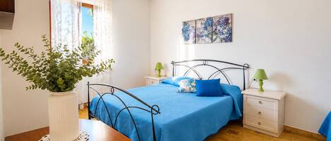 A bedroom with a bed, bedside tables, a nightstand, a potted plant, and a painting on the wall.