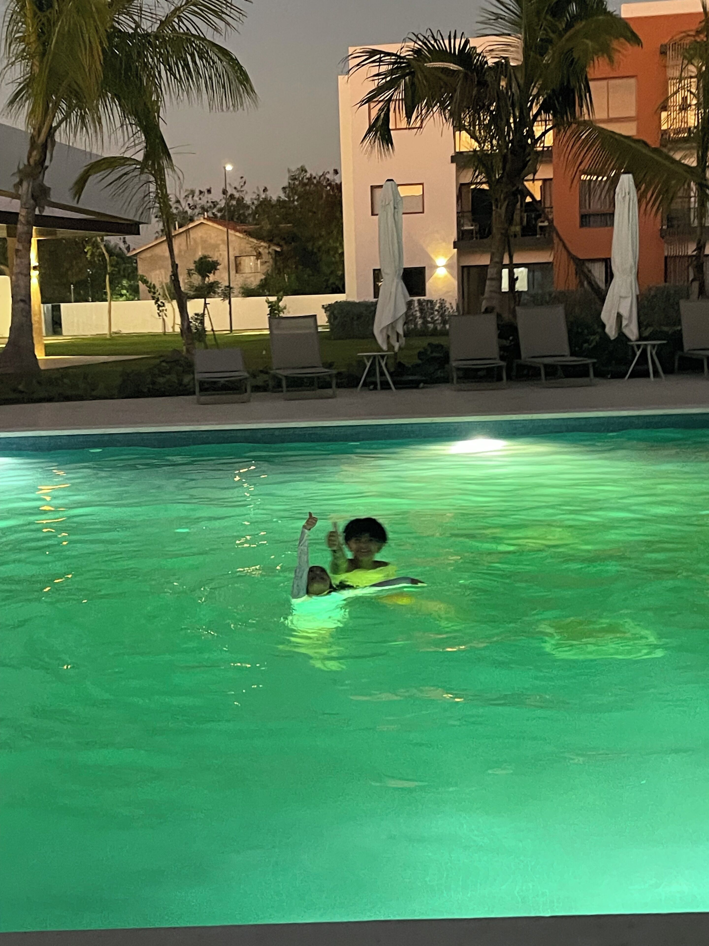 Ein Schwimmbecken mit zwei Personen im Wasser, umgeben von Liegestühlen und Sonnenschirmen, mit einem Gebäude im Hintergrund.