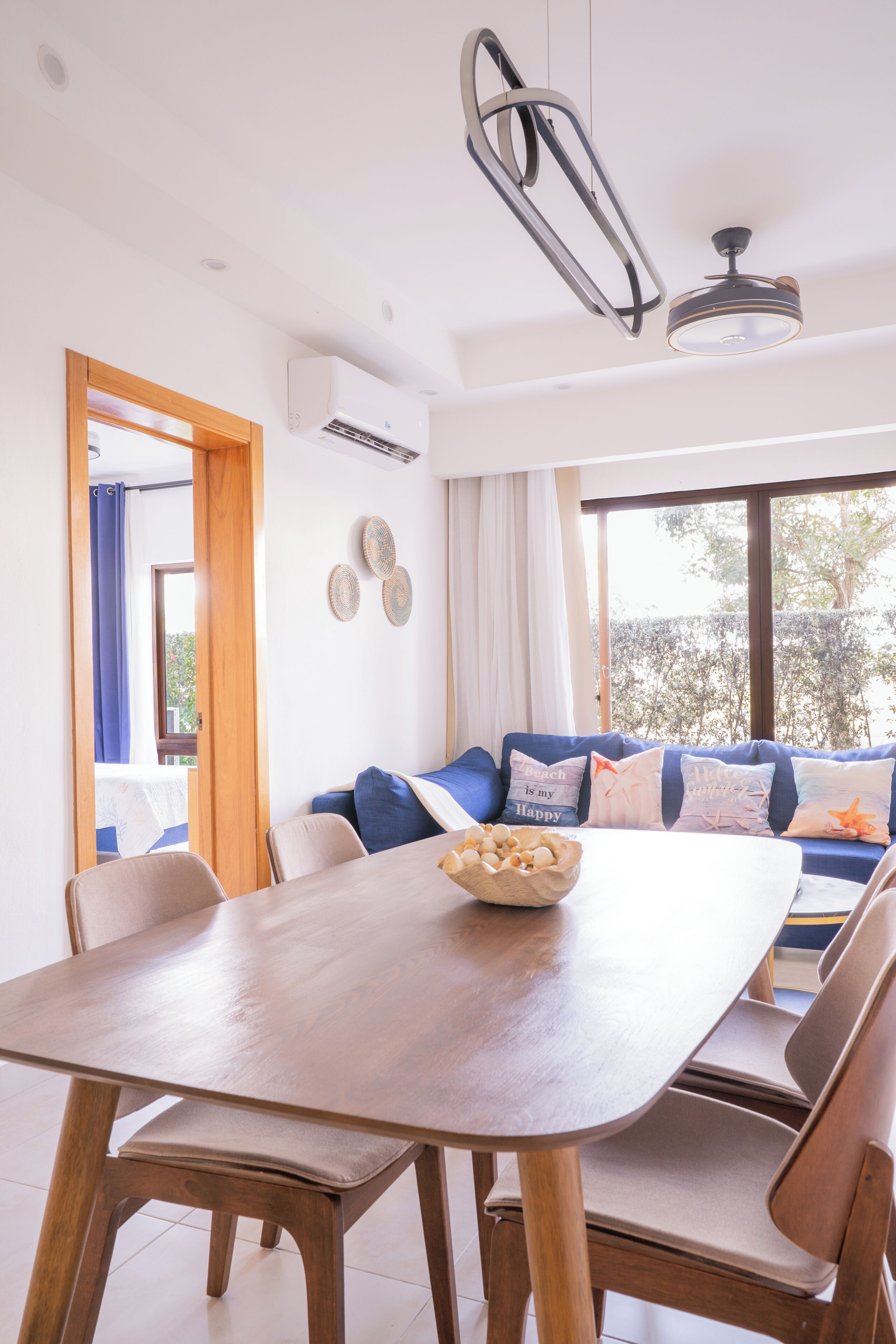 A modern dining area with a wooden table and chairs, a blue sofa with decorative pillows, and a view of the outdoors through large windows.