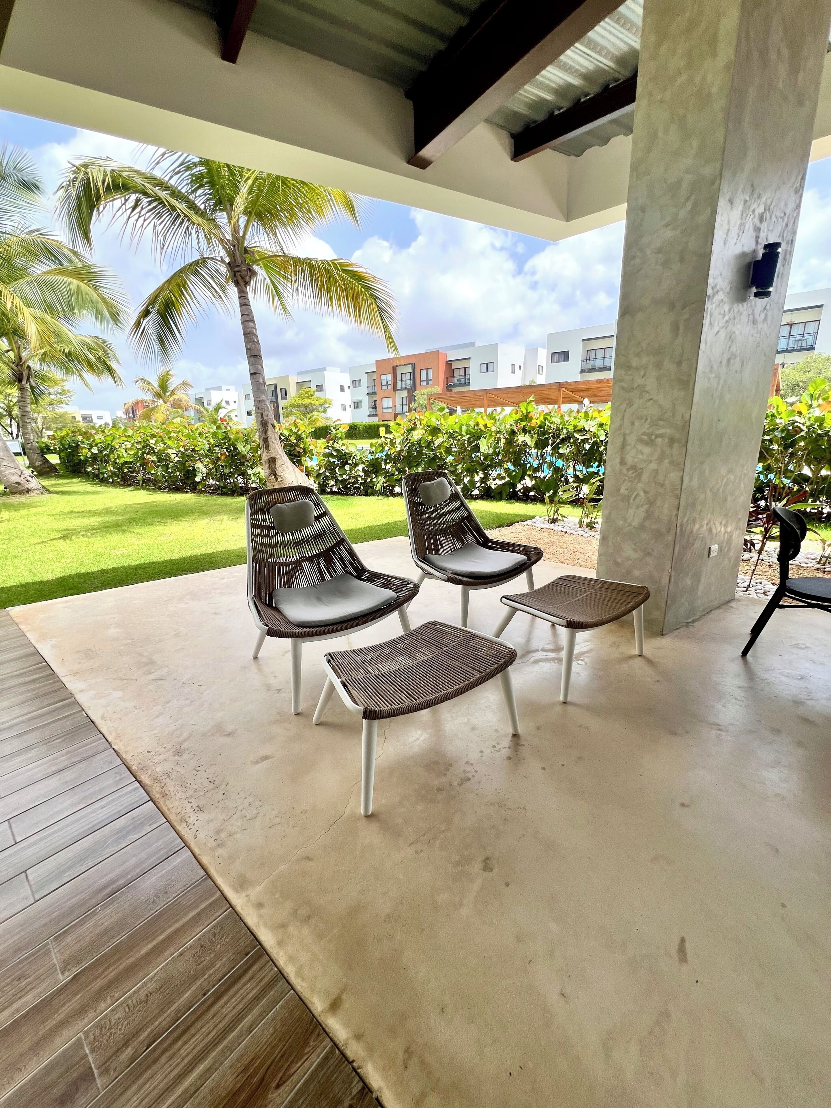 A covered outdoor seating area with chairs and a table, palm trees, and a view of buildings in the background.