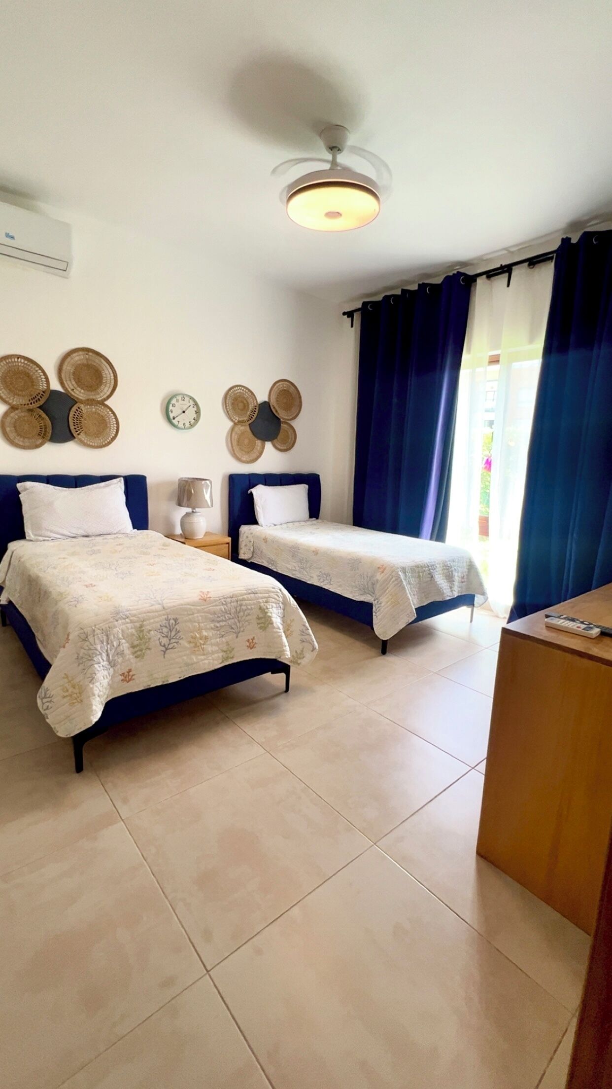 Ein Zimmer mit zwei Betten, einem Deckenventilator, einer Uhr und einem Fenster mit Vorhängen.