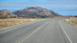 Una carretera serpentea por un paisaje árido y montañoso, con poca vegetación y una montaña al fondo.