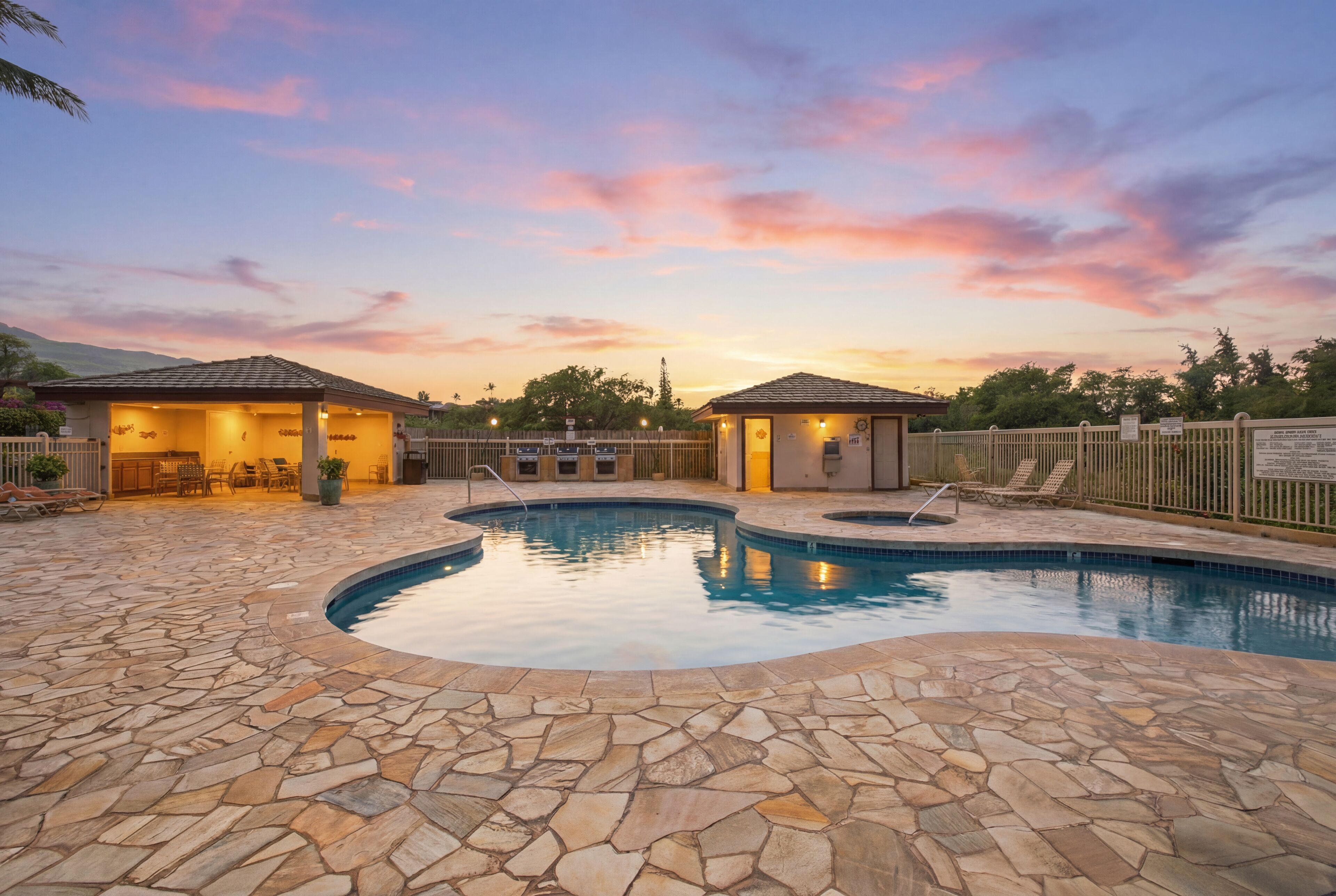 Una piscina con baldosas de piedra, dos pequeñas construcciones con techo y un área vallada con sillas.