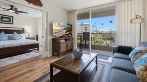 A bedroom with a bed, a TV, a coffee table, a sofa, and a balcony with a view of palm trees and buildings. - Brand-New Interior, 5-Min Walk to Beach, 2 Baths (Kihei)