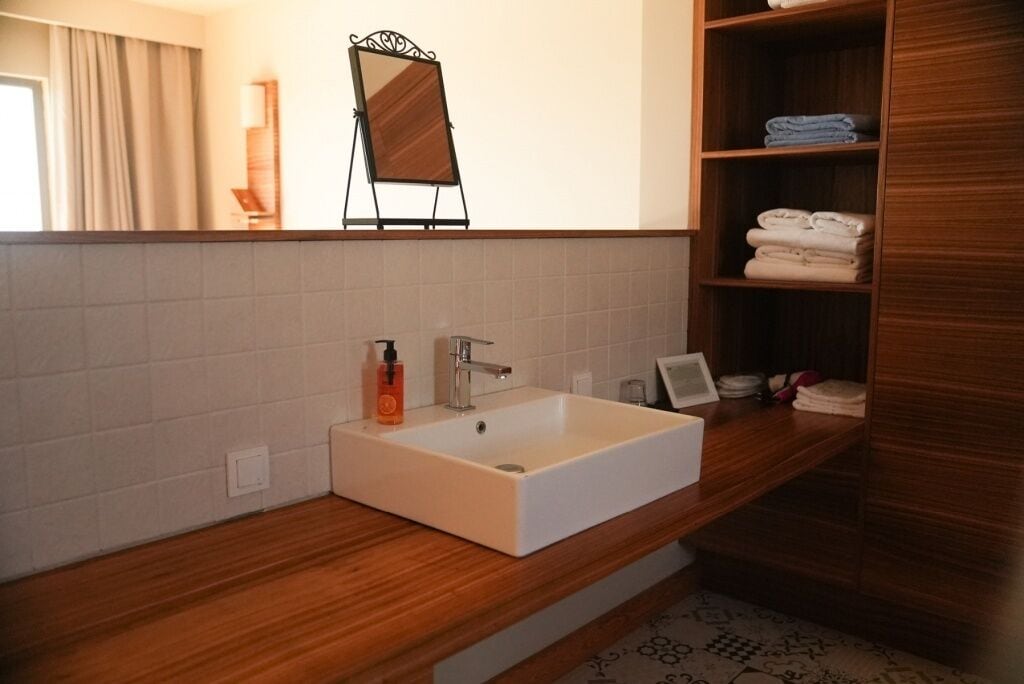 A bathroom with a white sink, wooden vanity, a mirror, and a towel shelf.