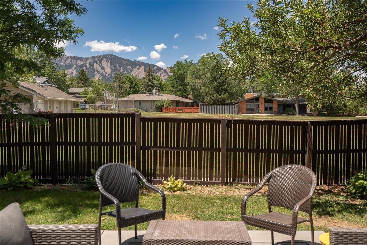 A backyard with a wooden fence, two wicker chairs, a small table, and a view of houses and mountains.