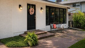 Un bâtiment blanc avec une porte noire, deux lampes fixées au mur, un banc avec des coussins et une plante en pot.
