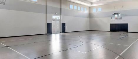 A spacious indoor basketball court with a high ceiling, multiple windows, and a basketball hoop.