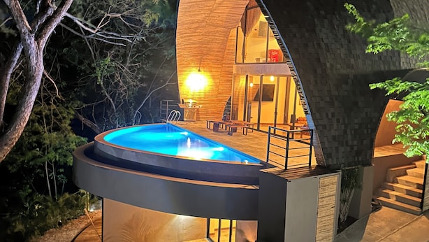 Een modern, gebogen gebouw met een hottub en een terras, omgeven door bomen.