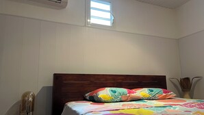 Une chambre à coucher avec une tête de lit en bois, une couvre-lit colorée, une petite fenêtre et un appareil de climatisation.