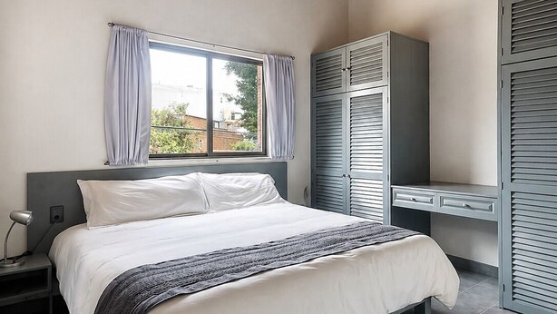 Ein Schlafzimmer mit Bett, Schreibtisch, Kleiderschrank, einem Fenster mit Vorhängen und einer Pendelleuchte.