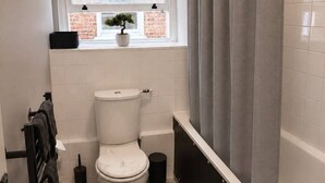 Une salle de bain avec des toilettes, une fenêtre avec une plante, une baignoire avec un rideau de douche gris et un tapis noir sur le sol.
