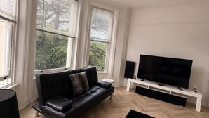 Un salon moderne avec un canapé en cuir noir, un téléviseur à écran plat et une grande fenêtre équipée de stores.