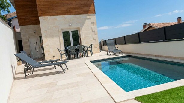 Una piscina amb gandules i una taula, un edifici de pedra amb teulada de fusta, i una tanca negra.