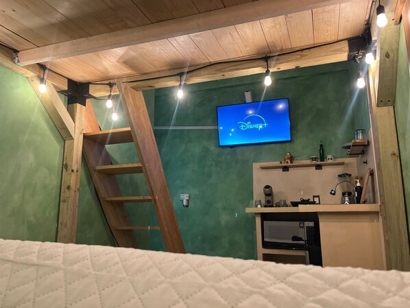 Ein Zimmer mit Holzboden, einem Fernseher, der Disney+ anzeigt, einer Leiter, einer Küchenzeile und einem Bett mit weißer Decke.