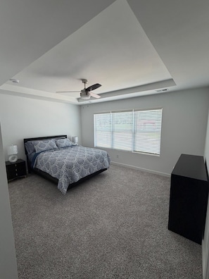 Une chambre à coucher avec un lit, un ventilateur de plafond, une fenêtre avec des stores, une table de chevet et un haut-parleur noir.
