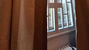 Ein Fenster mit braunen Vorhängen, teilweise geöffnet, das einen Blick auf ein Mehrstöckige Gebäude mit Balkonen freigibt.