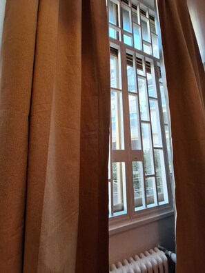 Ein Fenster mit braunen Vorhängen, teilweise geöffnet, das einen Blick auf ein Mehrstöckige Gebäude mit Balkonen freigibt.