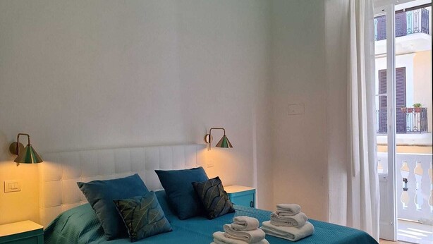 Ein Schlafzimmer mit einer türkisfarbenen Bettdecke, einem Nachttisch, einer Lampe und Blick auf den Balkon.