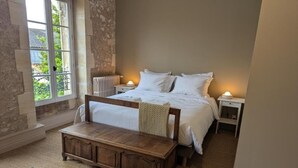 Un dormitorio con una cama grande, mesitas de noche, un banco de madera, un tapete estampado y vistas a través de una ventana.