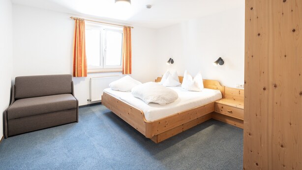A bedroom with a bed, a sofa, a nightstand, a wooden wardrobe, and a window with curtains. - Holiday Apartment in St. Anton (Sankt Anton am Arlberg)