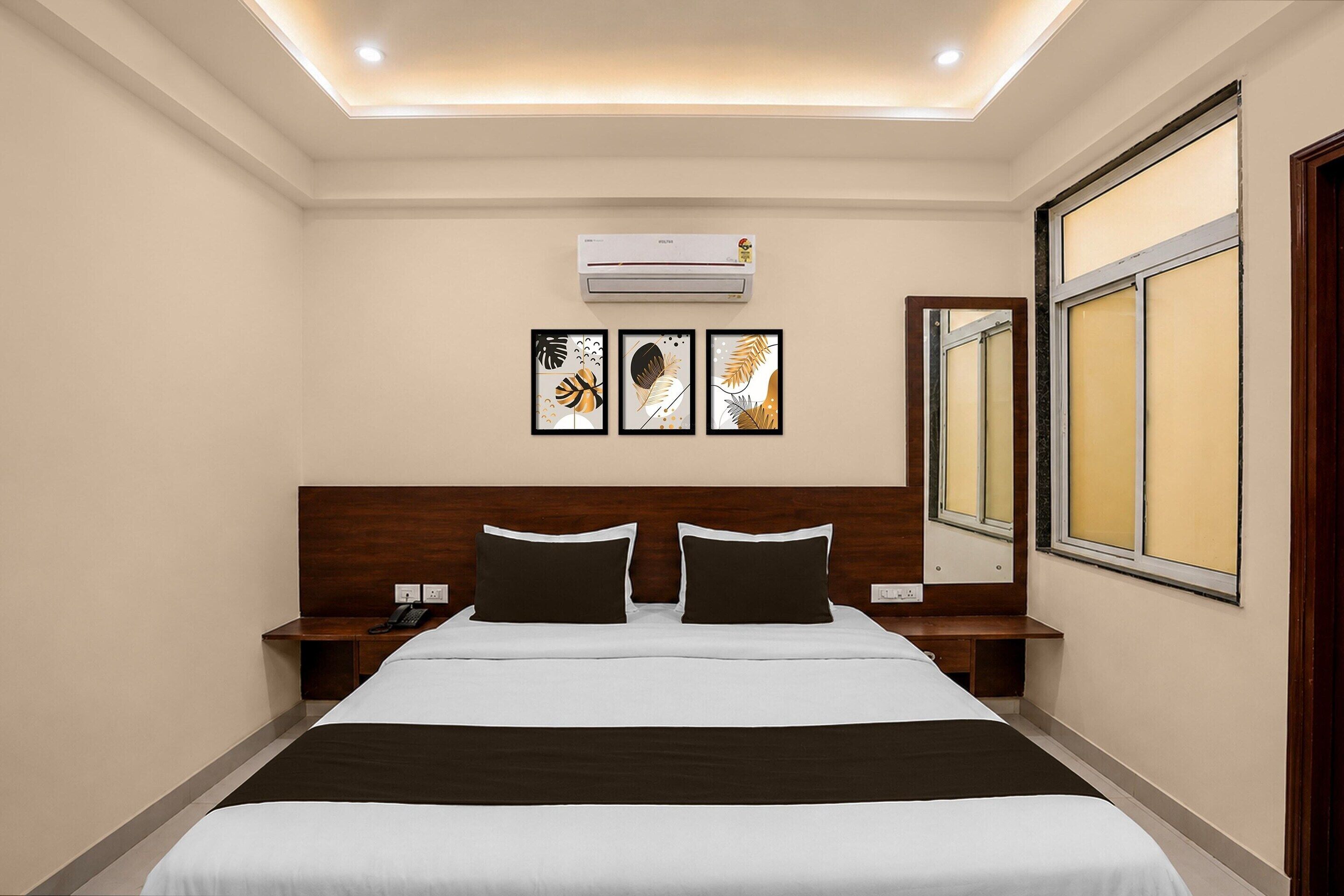 A hotel room with a large bed, two bedside tables, an air conditioning unit, and three framed artworks on the wall.
