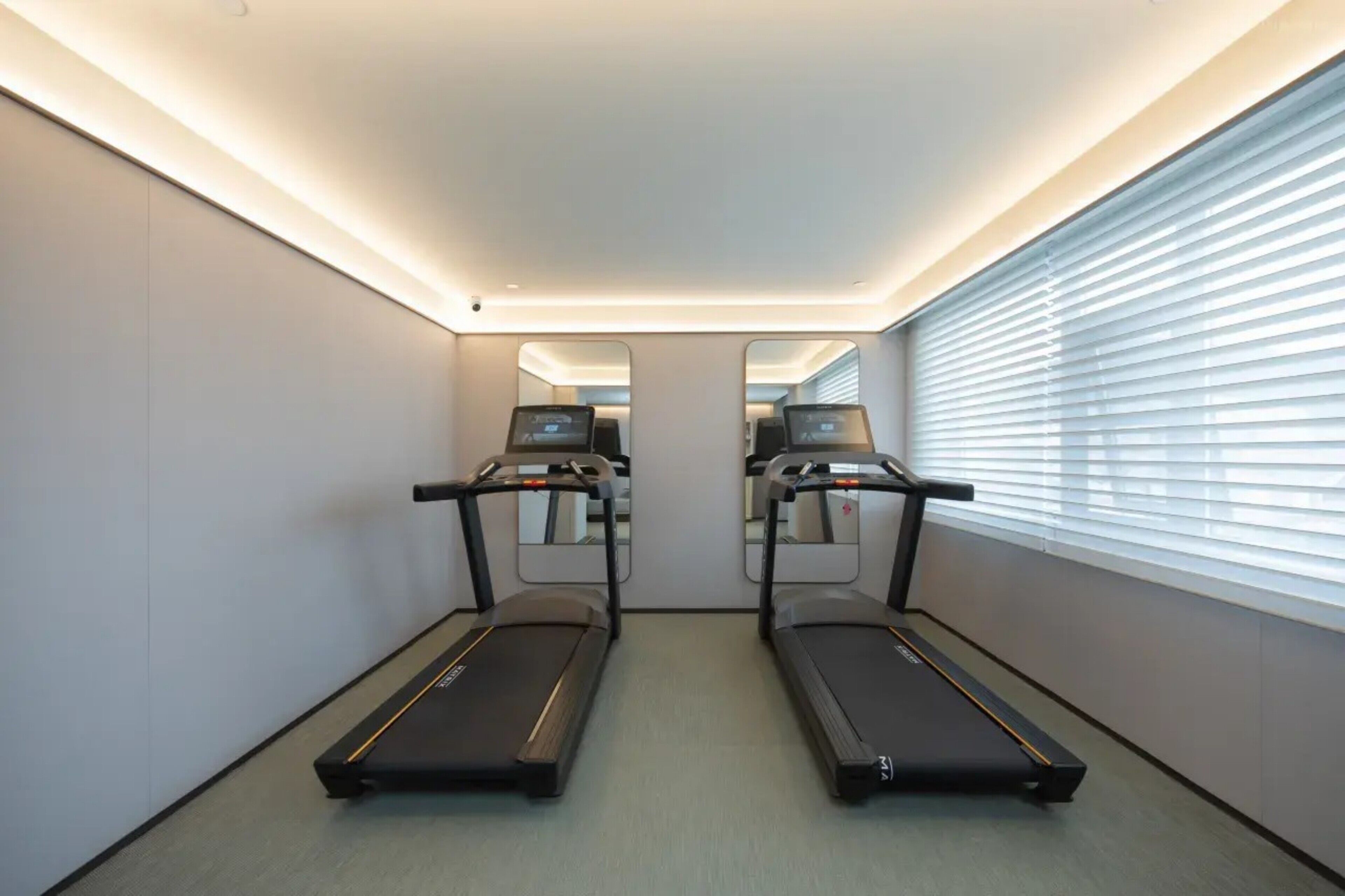 A room with two treadmills, a large window with blinds, and a mirror on the wall.