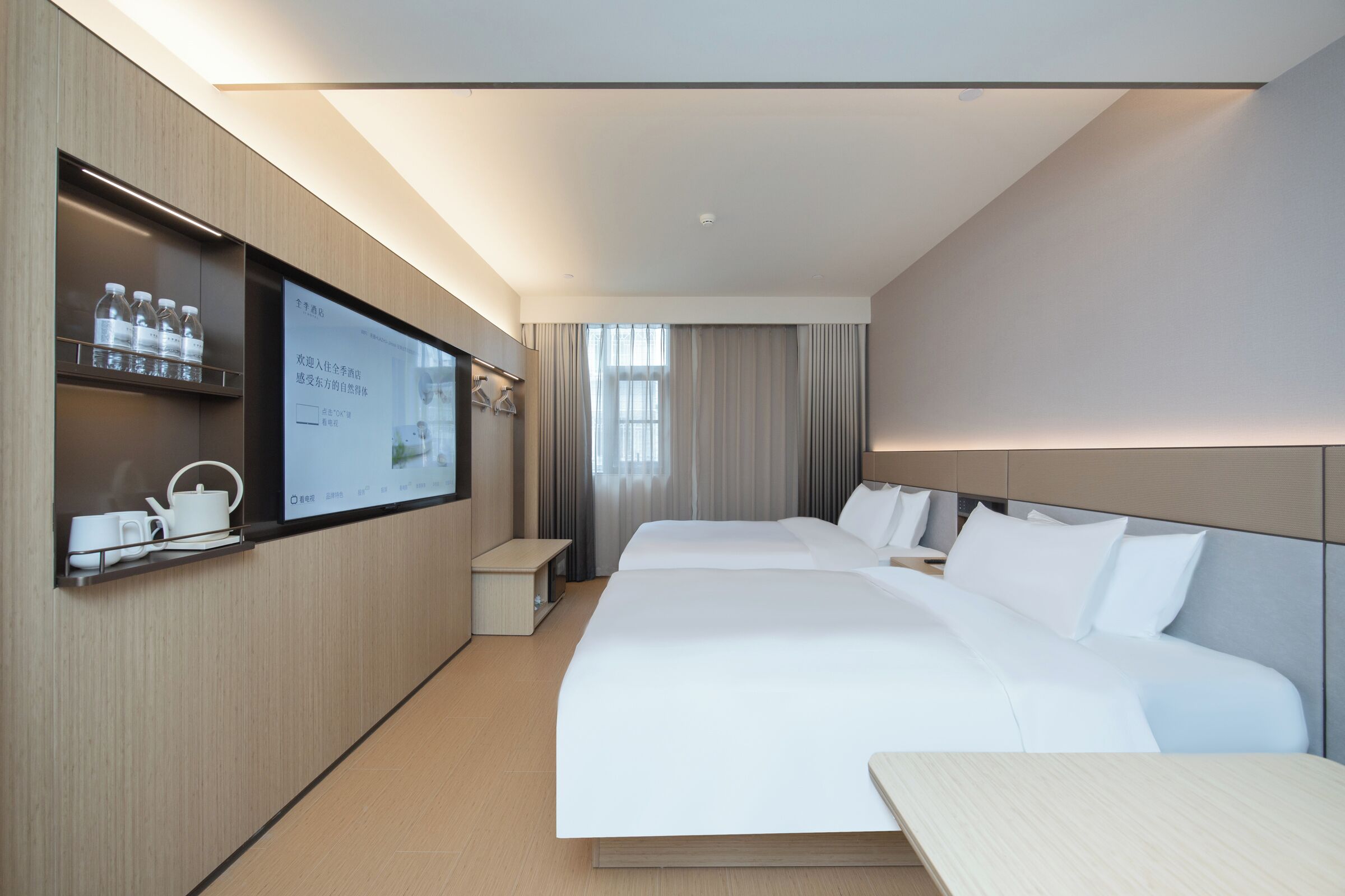A hotel room with a large bed, a bedside table, a TV mounted on the wall, and a shelf with water bottles and a tea set.