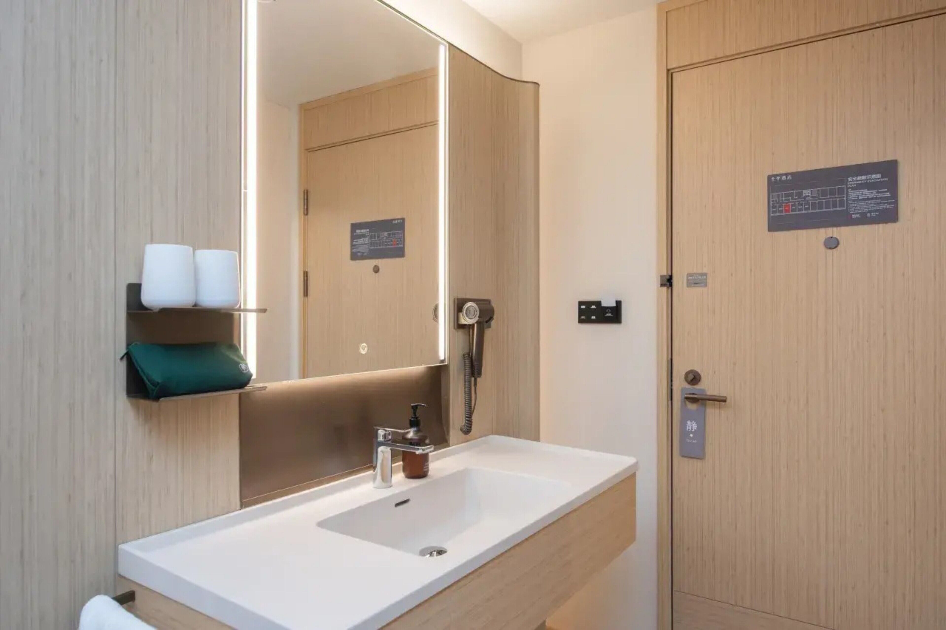 A modern bathroom with a large mirror, a sink, a towel dispenser, and a door with a sign.