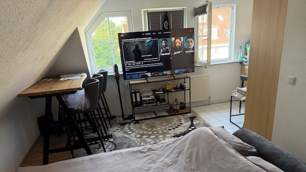 A room with a bed, a desk with a computer, a chair, a radiator, a shelf with items, and a window with a view of buildings.