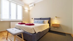 Un dormitorio con cama, una banca, una ventana y un estante fijado a la pared.