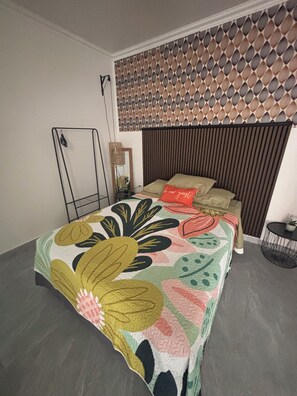 Ein Schlafzimmer mit einem gemusterten Kopfteil, einer blumigen Bettdecke, einem Nachttisch mit Lampe und einem kleinen Topfpflanzen.