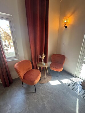 Una habitació amb dues cadires taronges, una taula rodona petita, un vas amb plantes seques, una cortina vermella i una làmpada fixada a la paret.