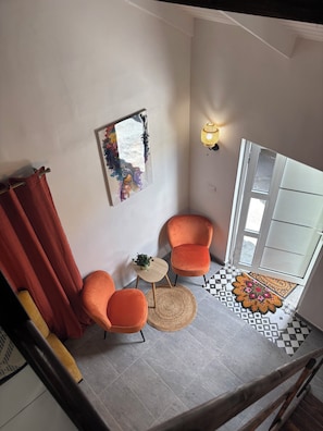 A corner room with two orange chairs, a small table with a plant, a colorful painting on the wall, and a patterned doormat.