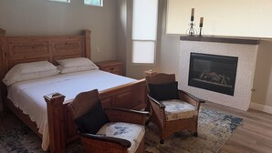Un dormitorio con una cama grande, dos sillones de mimbre, una chimenea y tres ventanas.