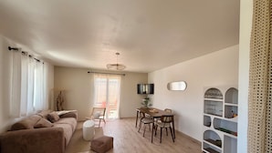 A living room with a sofa, a dining table with chairs, a TV mounted on the wall, and a large window with curtains. - La'PaillotteduBassin Vacation Rental (Le Teich)