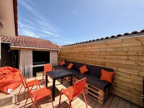 Une terrasse en bois avec une table noire, des chaises orange, un canapé en bois avec des coussins orange et un mur en bois.