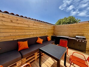 Une terrasse extérieure avec une plateforme en bois, une table noire, un canapé avec des coussins orange et un barbecue.