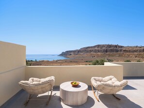 A balcony with two cushioned chairs and a round table, overlooking a coastal landscape with cliffs and the sea. - CASA 3 Lindos (Lindos)