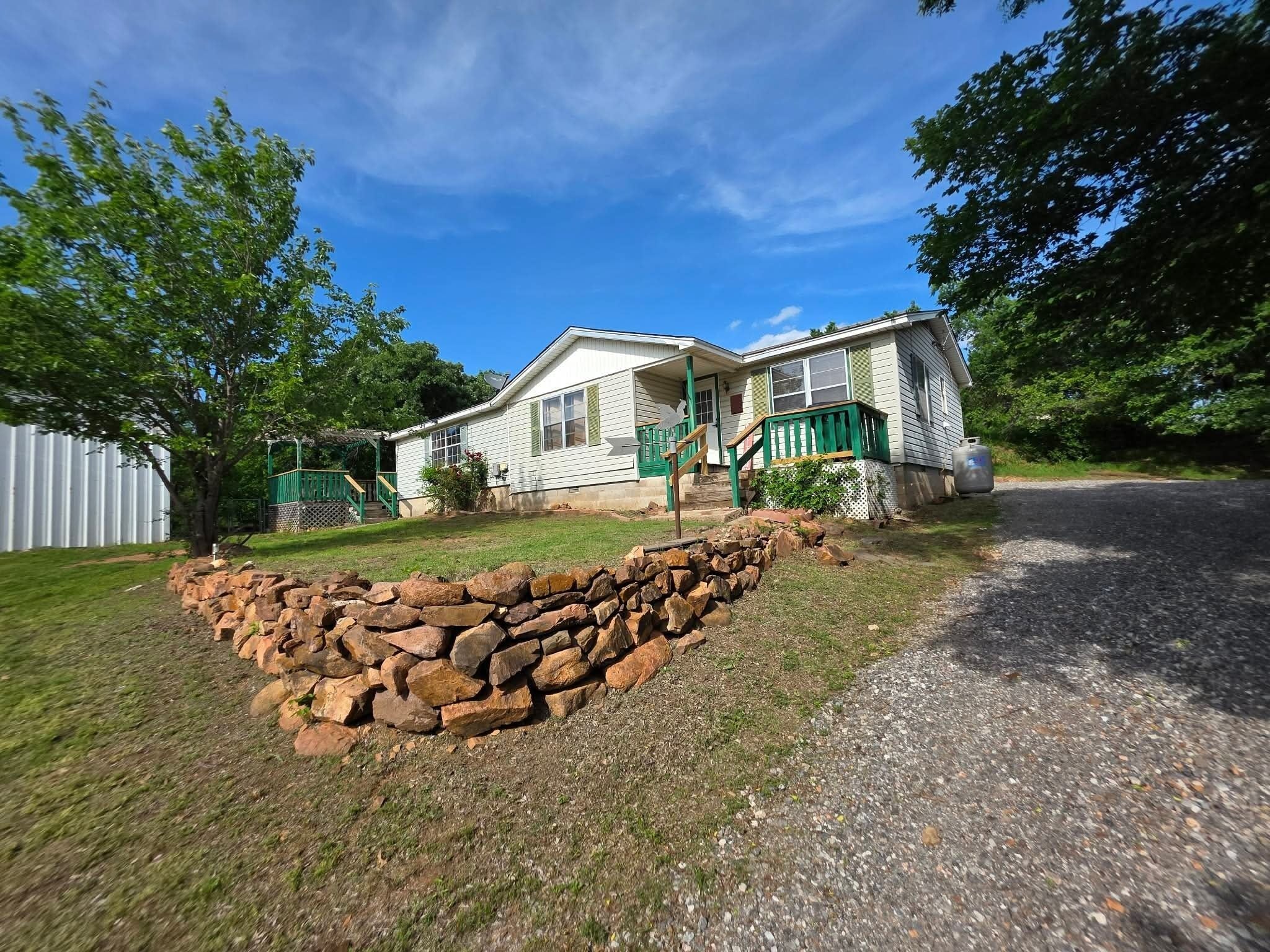 A single-story mobile home with a front porch, a stone retaining wall, and a gravel driveway.