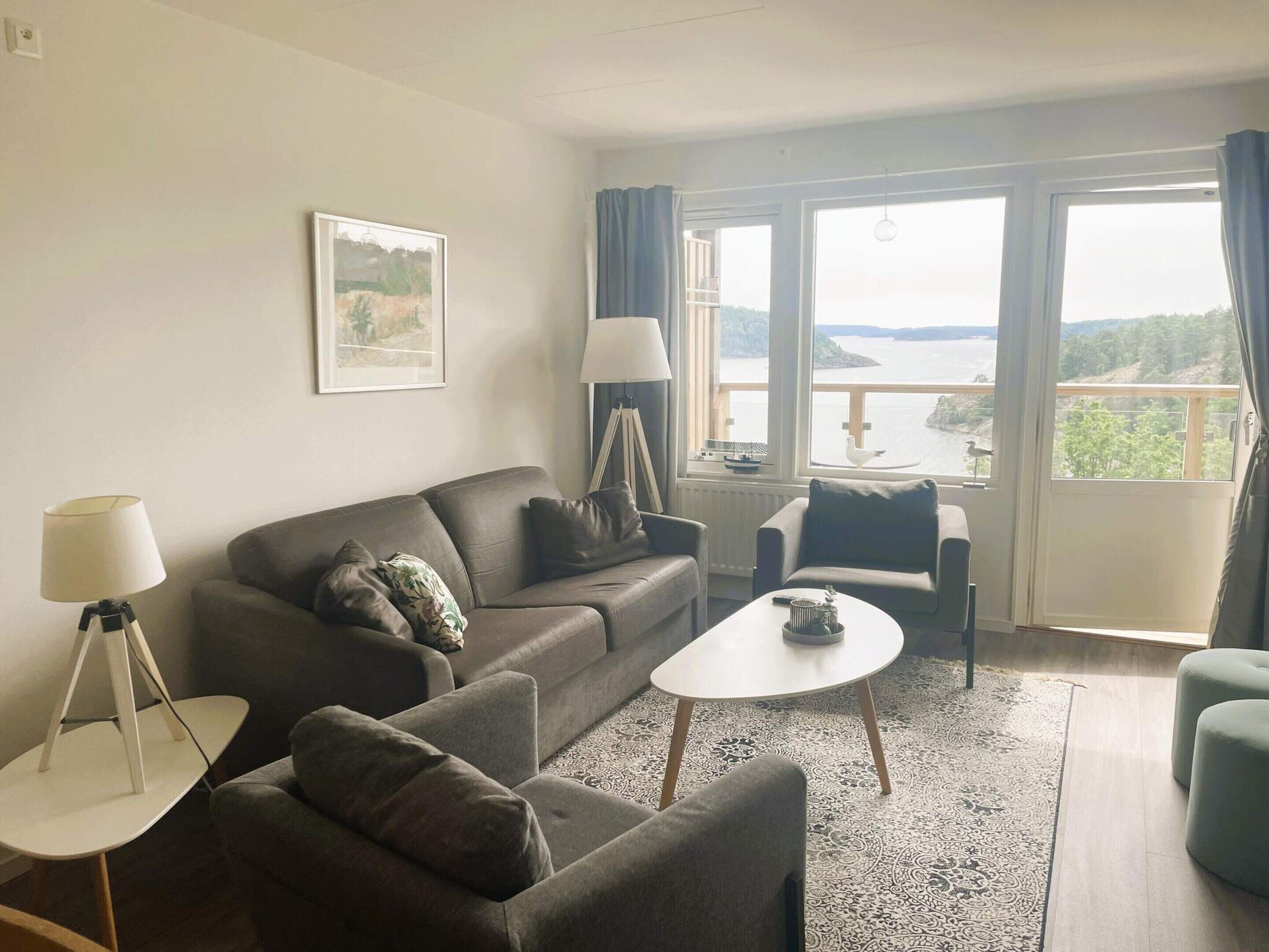 A living room with a sofa, armchair, coffee table, and a view of a body of water through large windows.