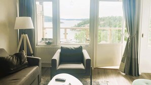 Habitación con sofá, sillón y una mesa con control remoto. Amplias ventanas con cortinas, vistas a un cuerpo de agua y vegetación.