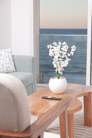 A room with a view of the ocean, a white vase with flowers on a wooden table, a remote control, and a light blue sofa with a patterned pillow. - sandra 17 (Tunis)