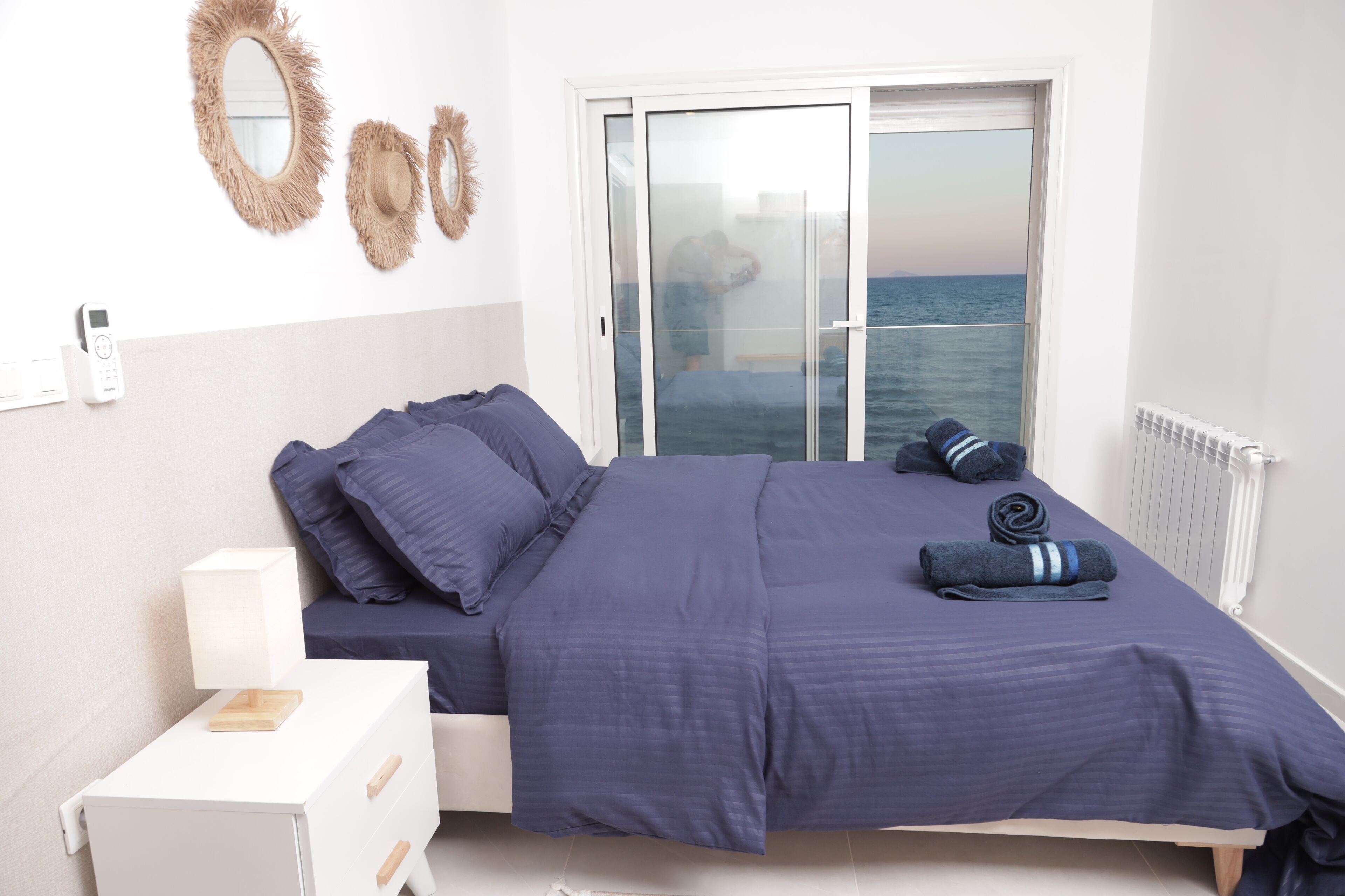 A bedroom with a bed, a nightstand, a lamp, and a view of the sea through a sliding door.