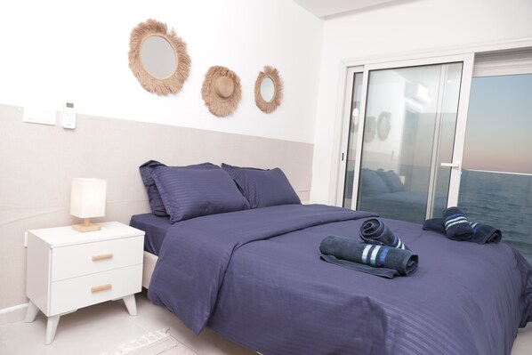 A bedroom with a bed, a nightstand, a lamp, and a view of the sea through a sliding door. - sandra 17 (Tunis)