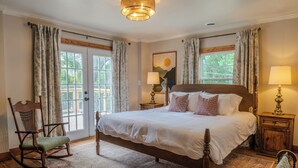 Une chambre à coucher avec un grand lit, deux tables de chevet, un fauteuil à bascule et une fenêtre avec des rideaux.